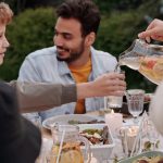Faceless female pouring fruit beverage into glass of boy in warm cloth while cheerful multiethnic family having dinner outdoors